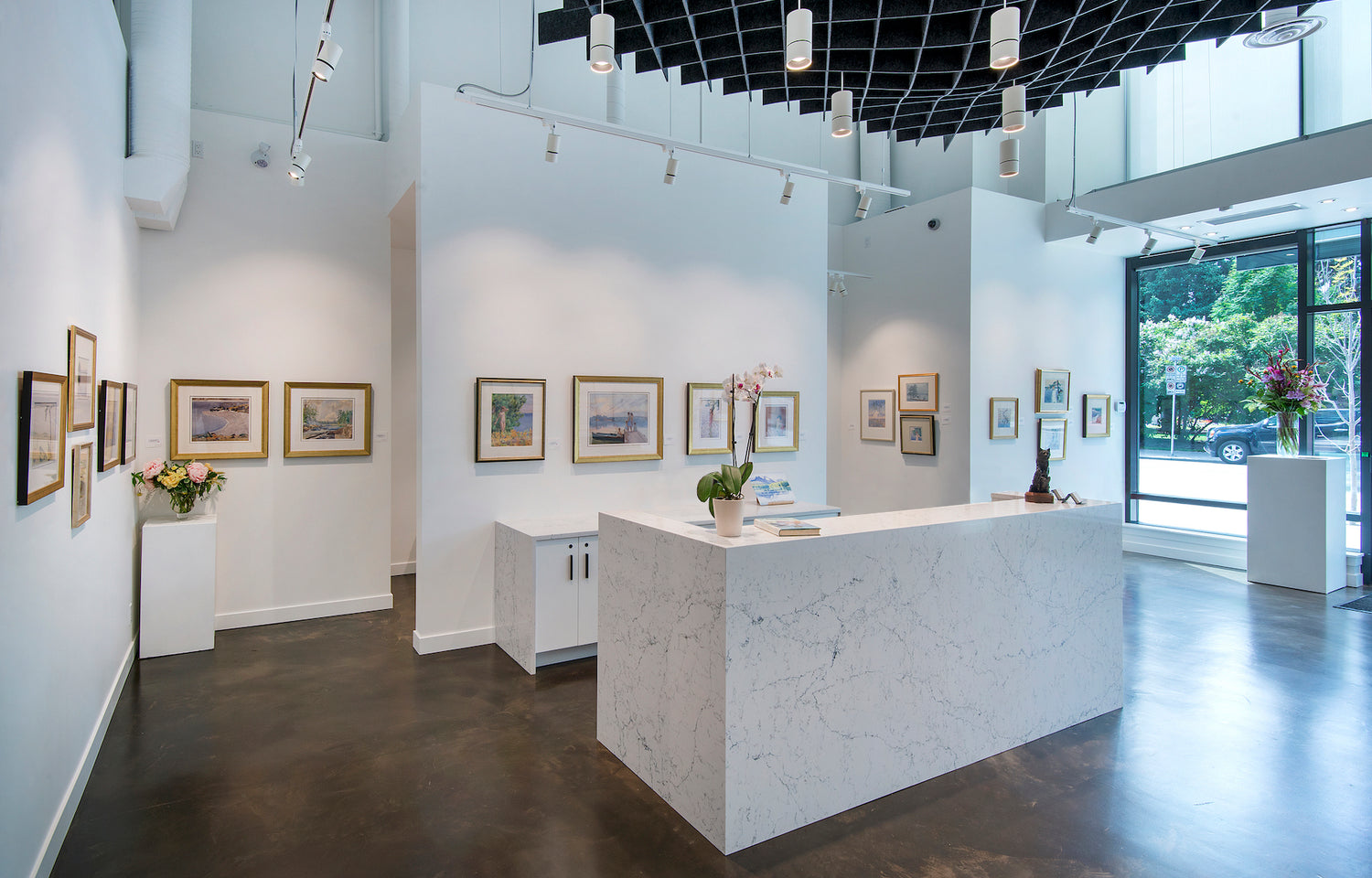 Photograph of a commercial art gallery with paintings hanging white walls, a bright window and bouquets of flowers.